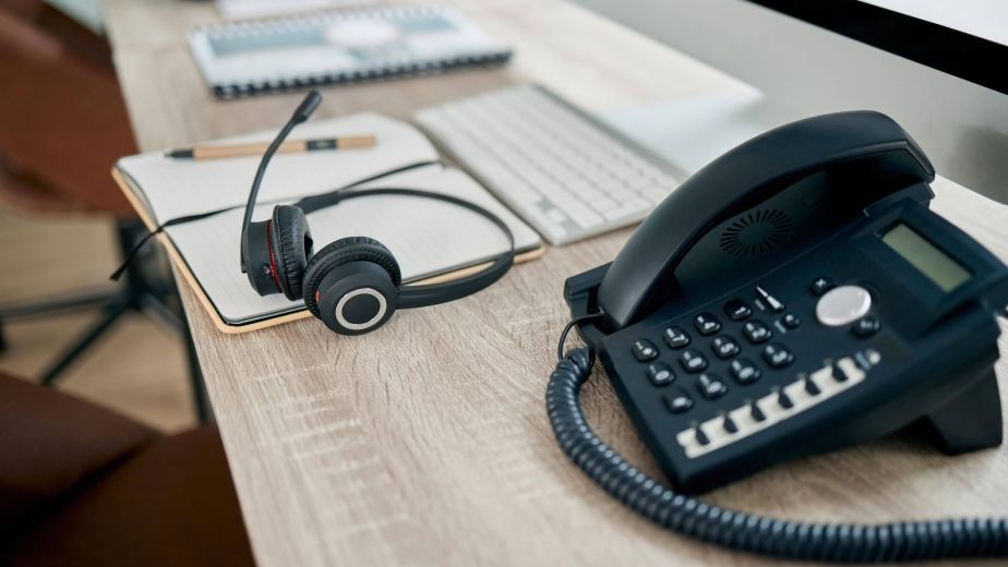 VoIP office phone and call centre headset on a wooden desk with a computer keyboard and notebook.